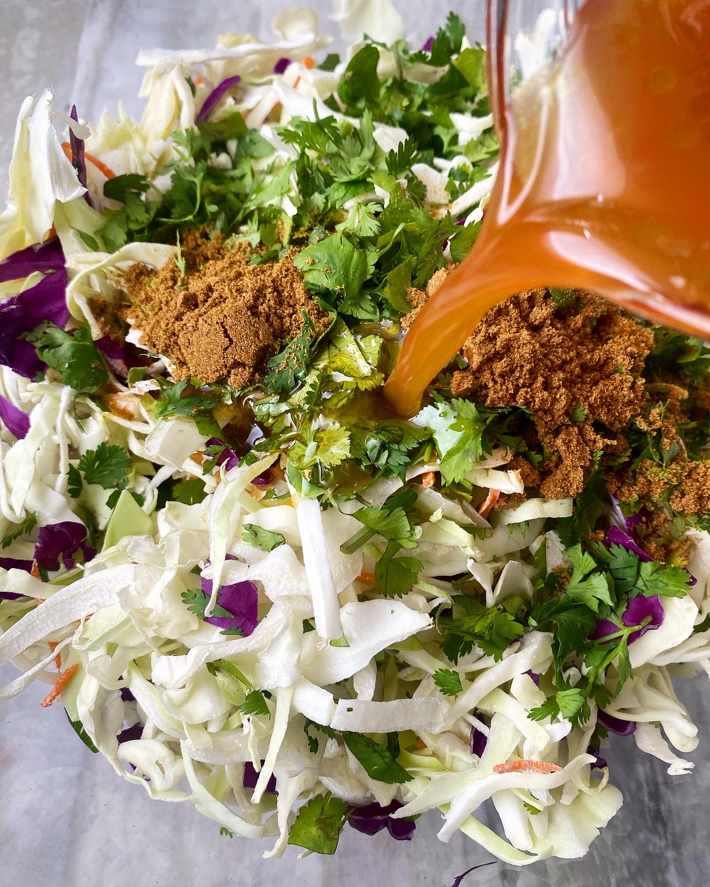 overhead shot of coleslaw in a glass bowl topped with fresh cilantro and cumin with a brown liquid mixture being poured in 