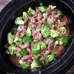 overhead shot of uncooked beef and broccoli in a black slow cooker with fresh broccoli added
