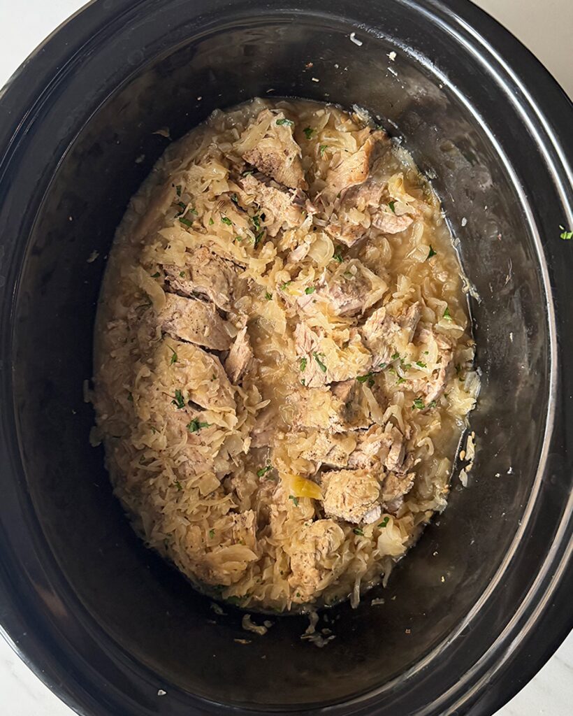 overhead shot of cooked Pork and Sauerkraut in a black slow cooker garnished with fresh parsley