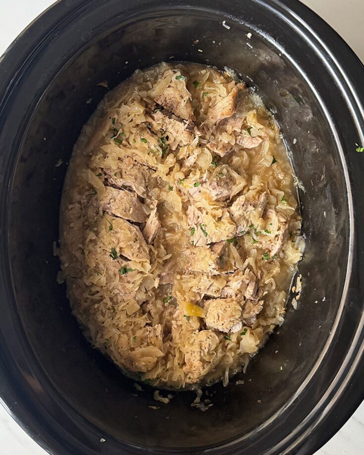 overhead shot of cooked Pork and Sauerkraut in a black slow cooker garnished with fresh parsley