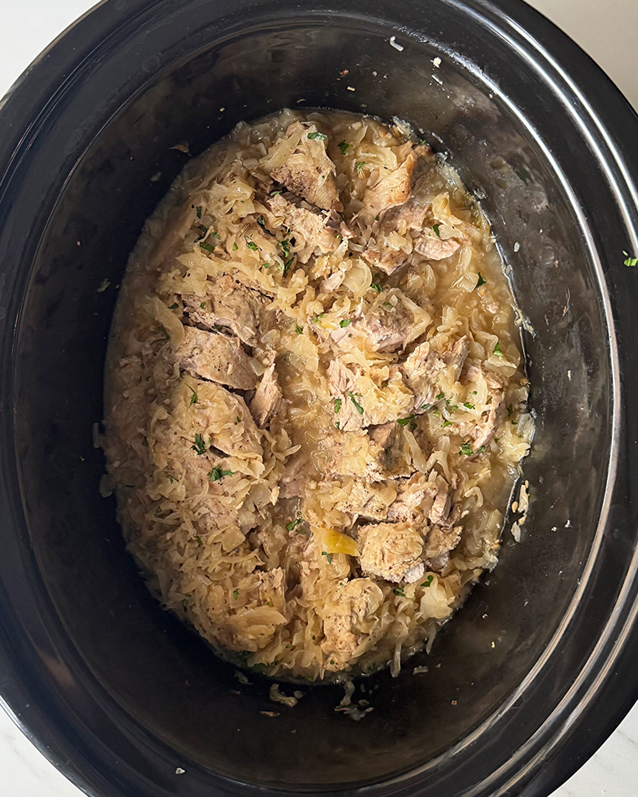 overhead shot of cooked Pork and Sauerkraut in a black slow cooker garnished with fresh parsley