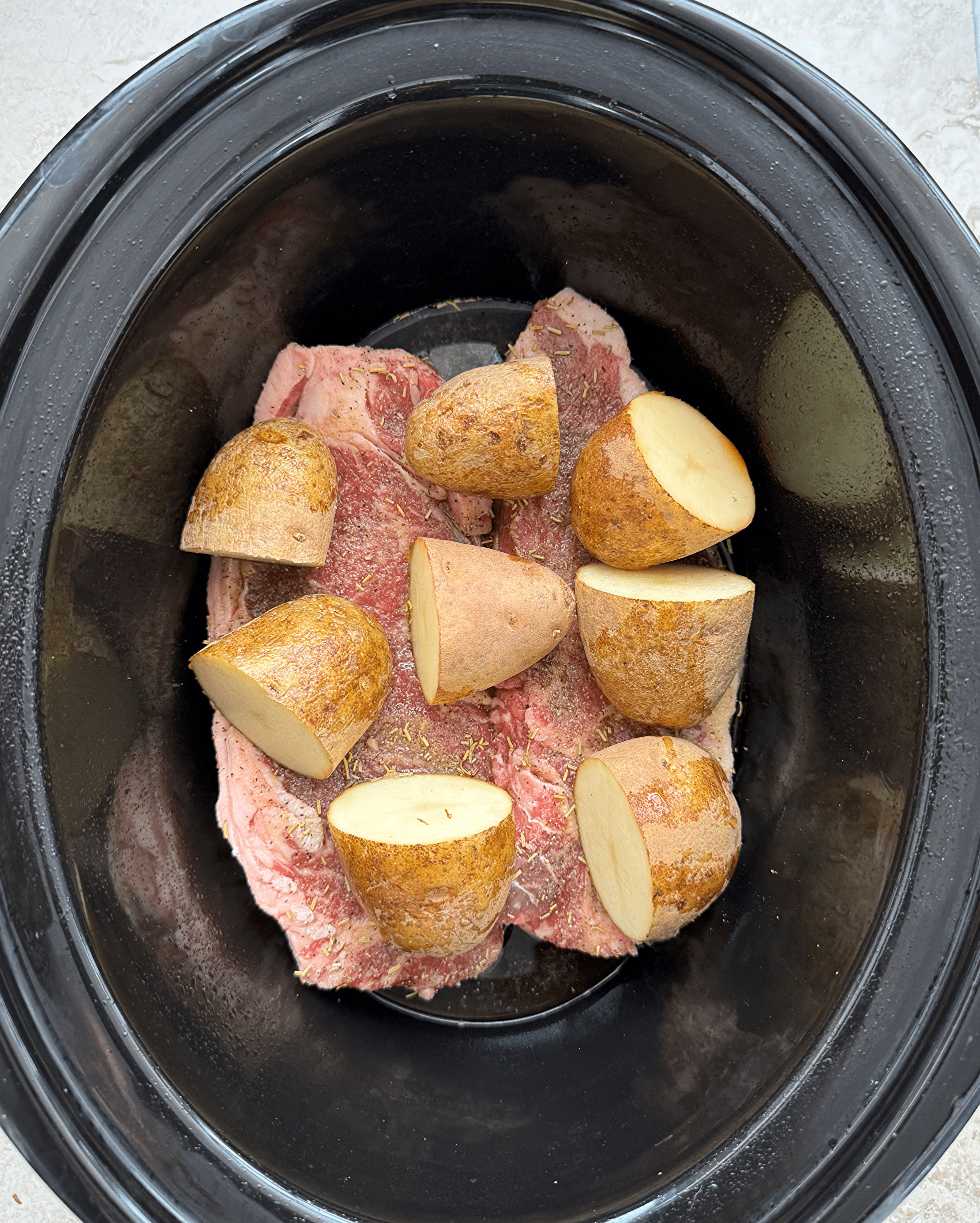 over head shot of uncooked steak and potatoes in a black slow cooker