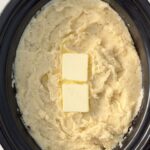 overhead shot of cooked mashed Potatoes in a black slow cooker with 2 tablespoons of butter on top