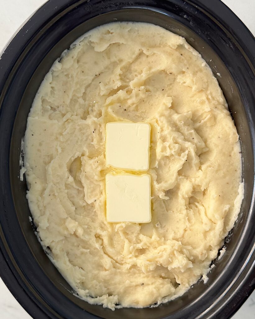 overhead shot of cooked mashed Potatoes in a black slow cooker with 2 tablespoons of butter on top