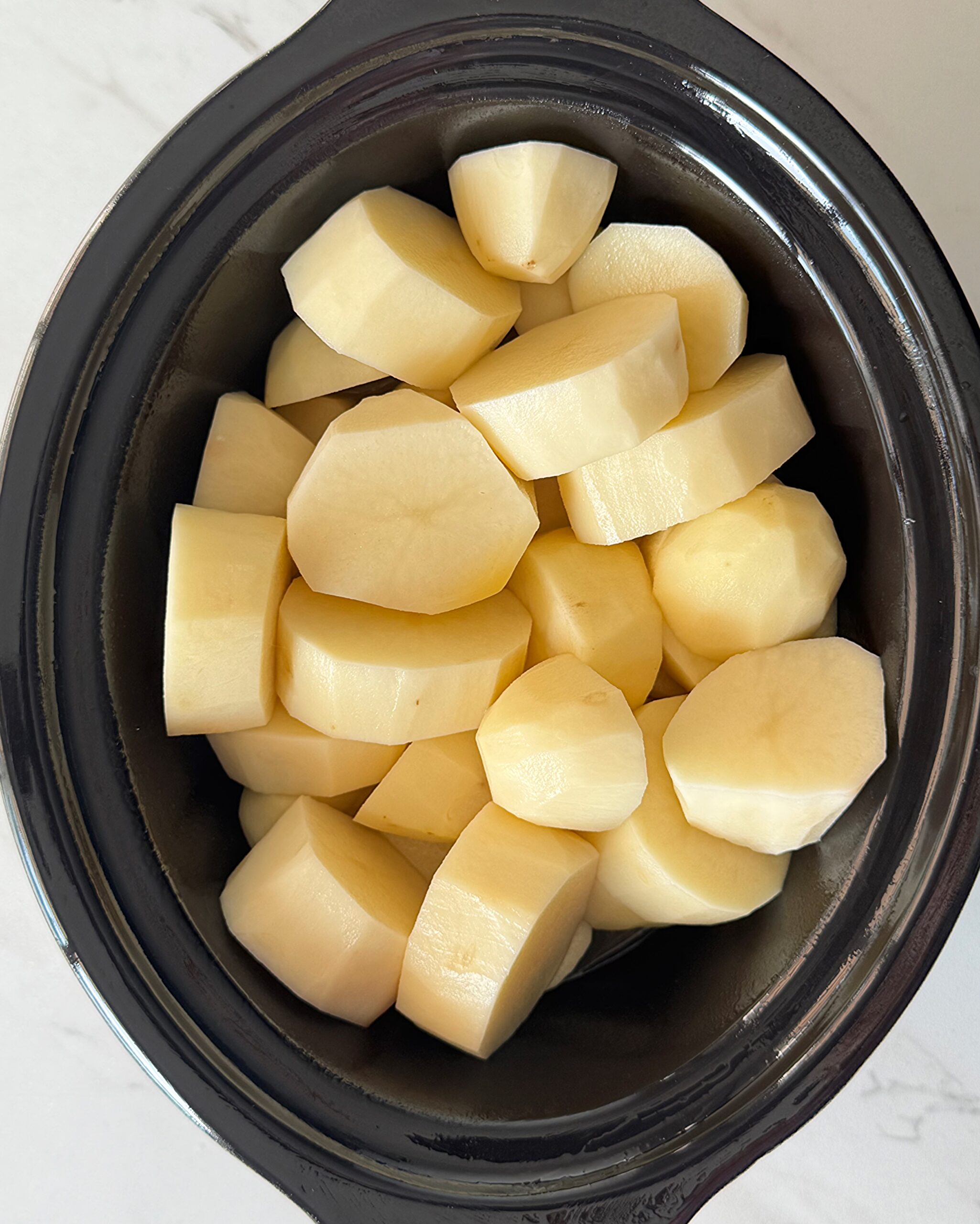 overhead shot of uncooked un-mashed Potatoes in a black slow cooker
