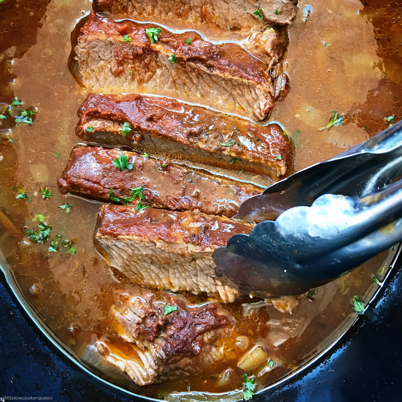 overhead close up shot of cooked, sliced bbq brisket in a black slow cooker with a silver tong taking a piece out