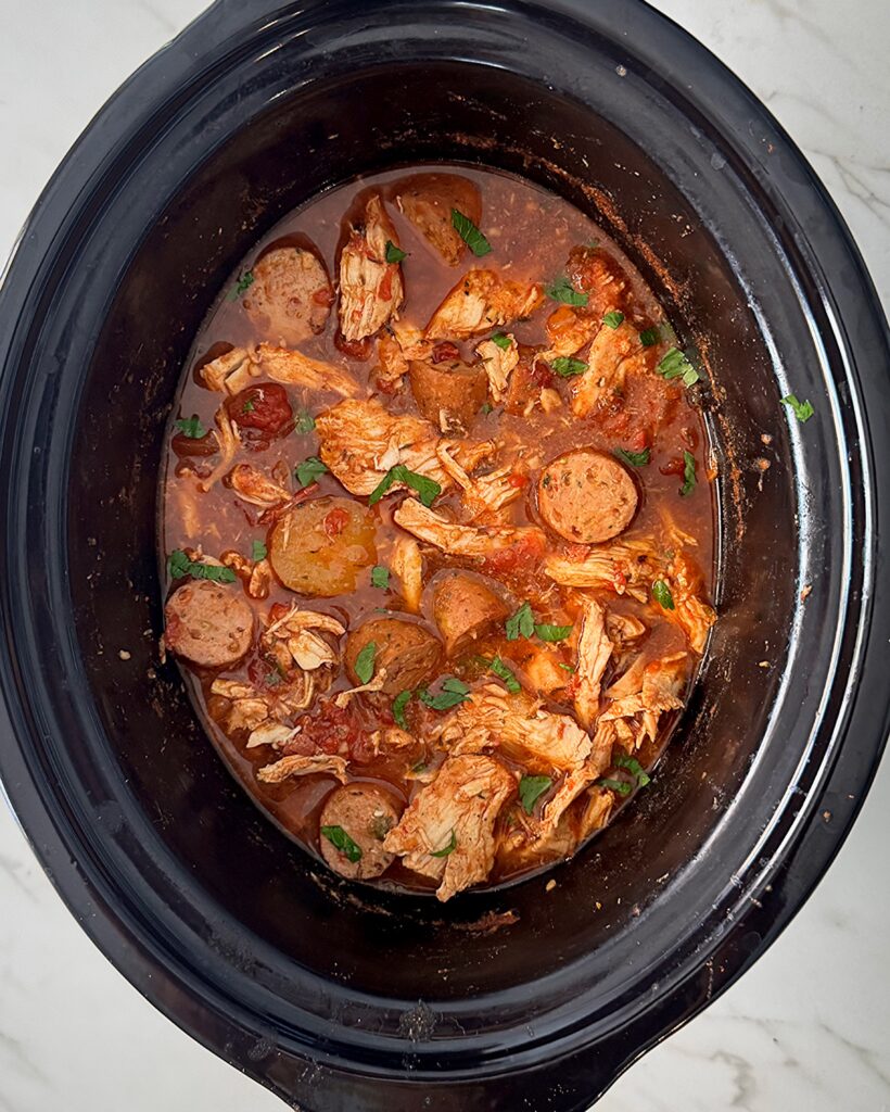 overhead shot of cooked spicy chicken and sausage in a black slow cooker garnished with fresh parsley