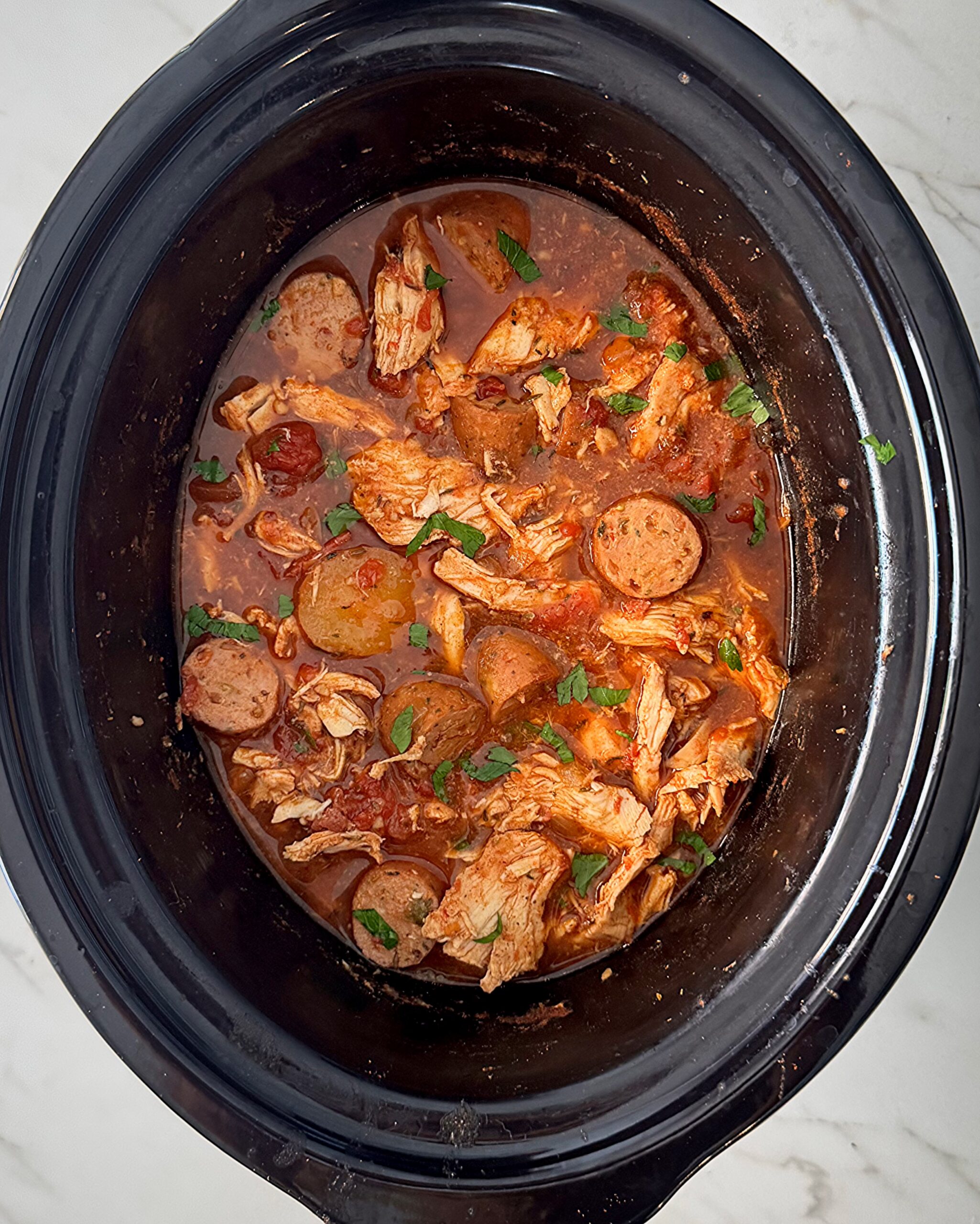 overhead shot of cooked spicy chicken and sausage in a black slow cooker garnished with fresh parsley