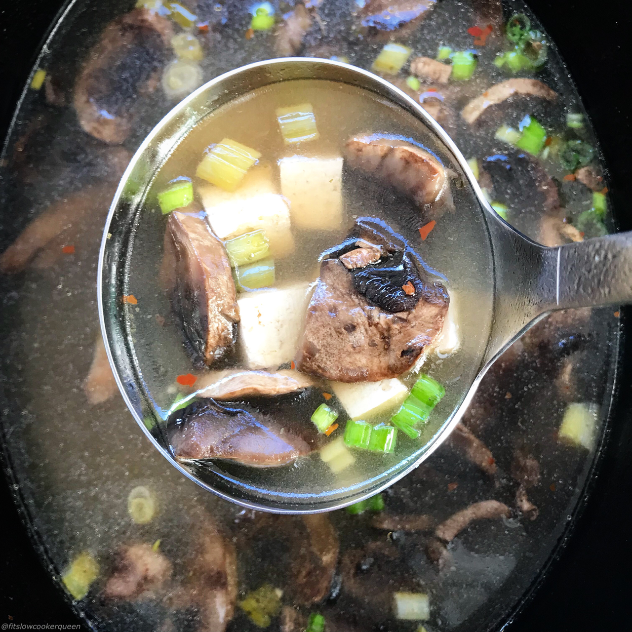 overhead shot of cooked tofu mushroom soup in a black slow cooker with a large silver spoon