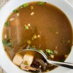 overhead shot of cooked tofu mushroom soup a white bowl (garnished with green onions) with a silver spoon