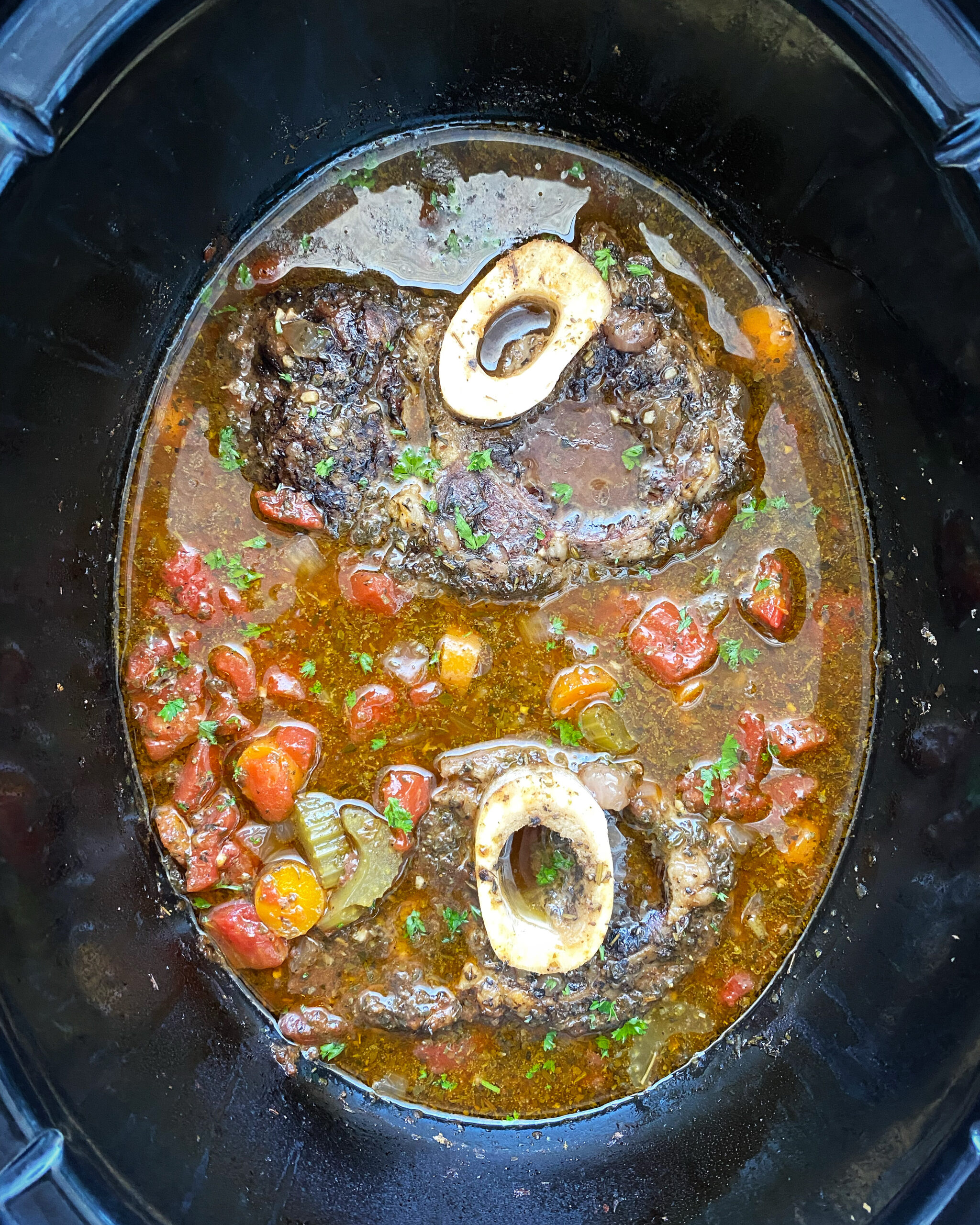 overhead shot of cooked crockpot beef osso buco in a black slow cooker garnished with fresh parsley