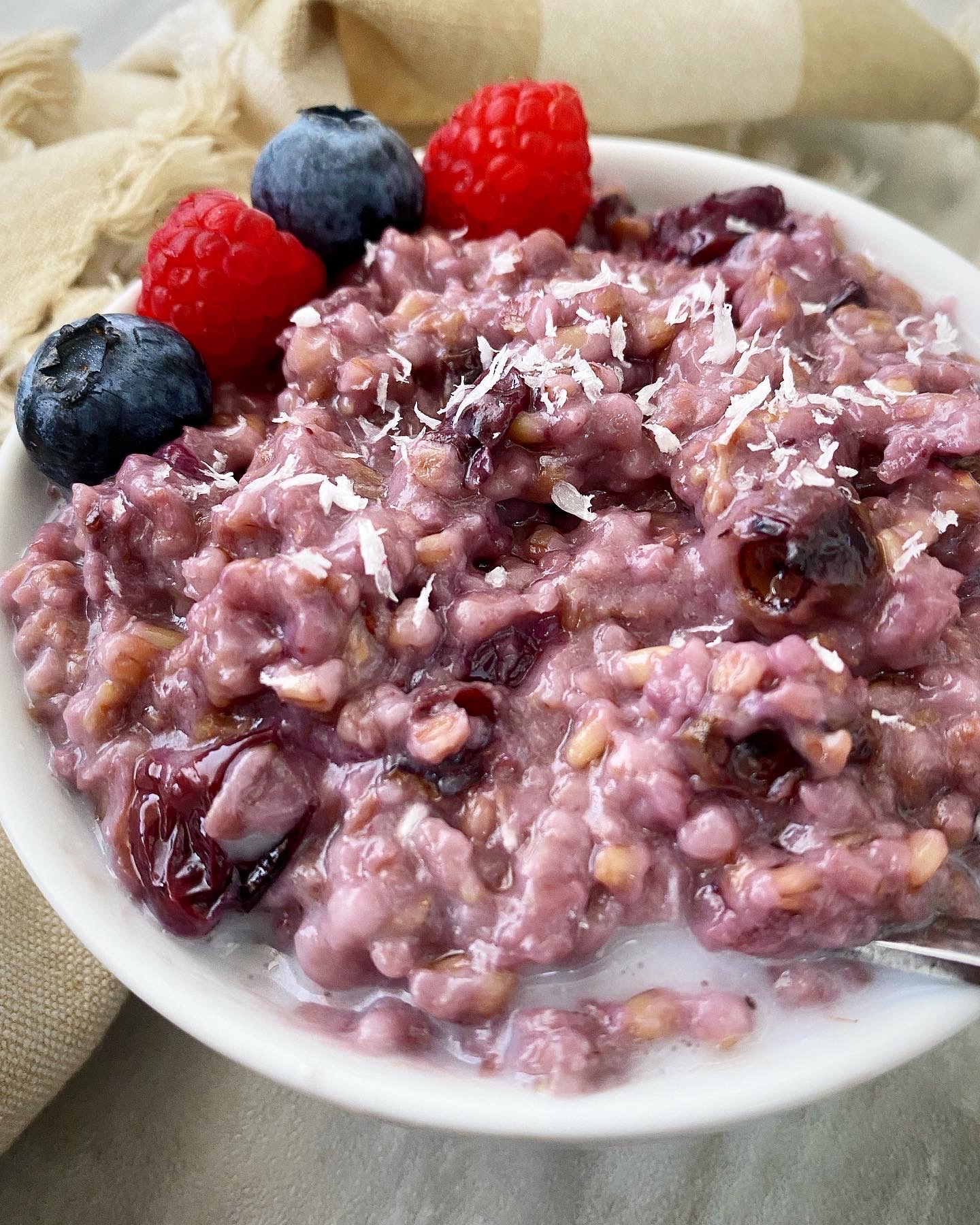 close up side view shot of berry oatmeal in white bowl garnished with fresh berries & shredded coconut