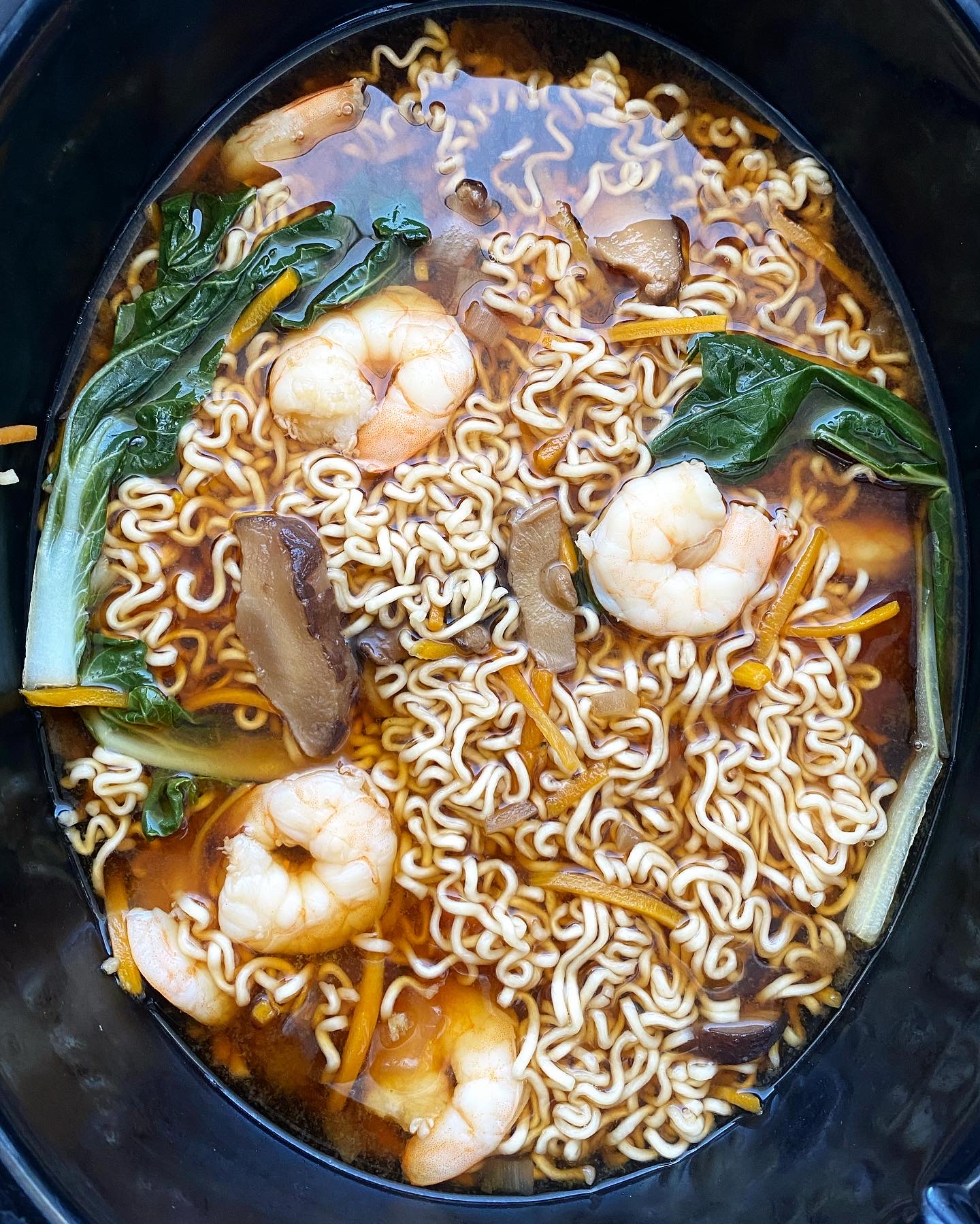 overhead shot of cooked soup with ramen, shrimp, and bok choy in a black slow cooker