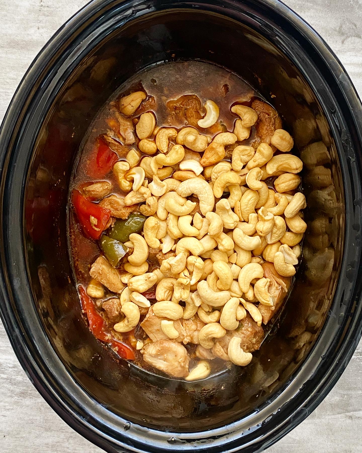 overhead shot of cooked chicken in a black slow cooker with cashews added