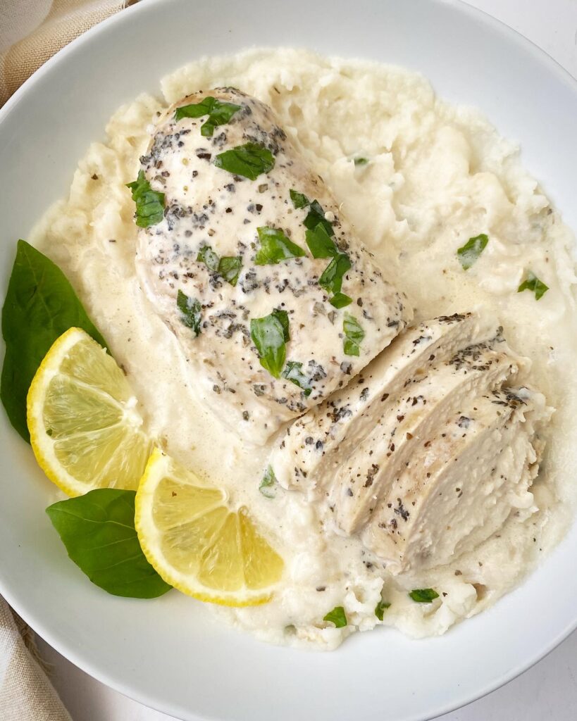 close up shot of lemon basil chicken sliced over a bed of mashed potatoes garnished with fresh basil and sliced lemon