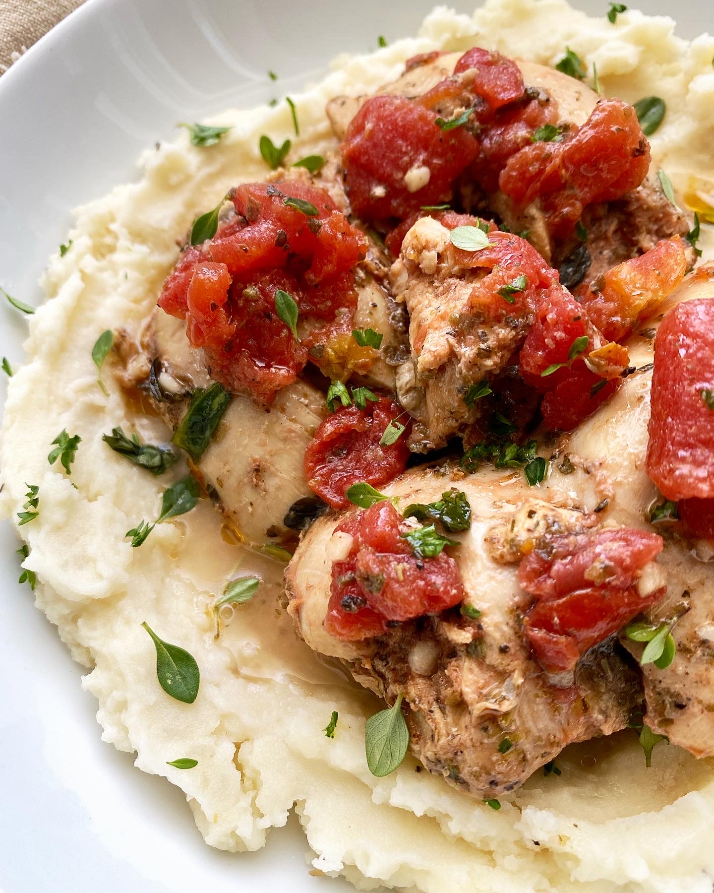 side view of tomato garlic chicken & herbs in a white bowl on top of mashed potatoes with fresh herbs on top