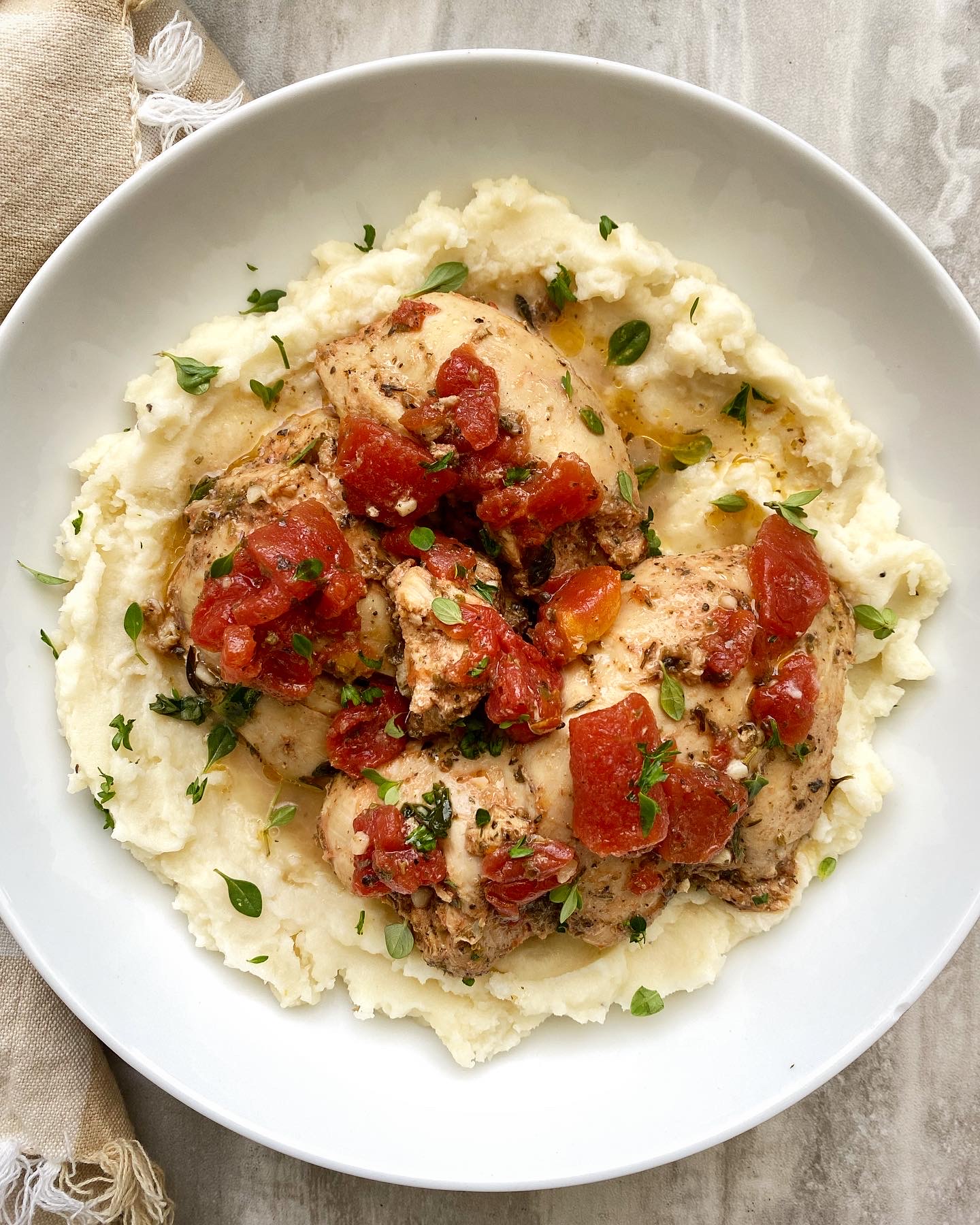 overhead shot of tomato garlic chicken & herbs in a white bowl on top of mashed potatoes with fresh herbs on top