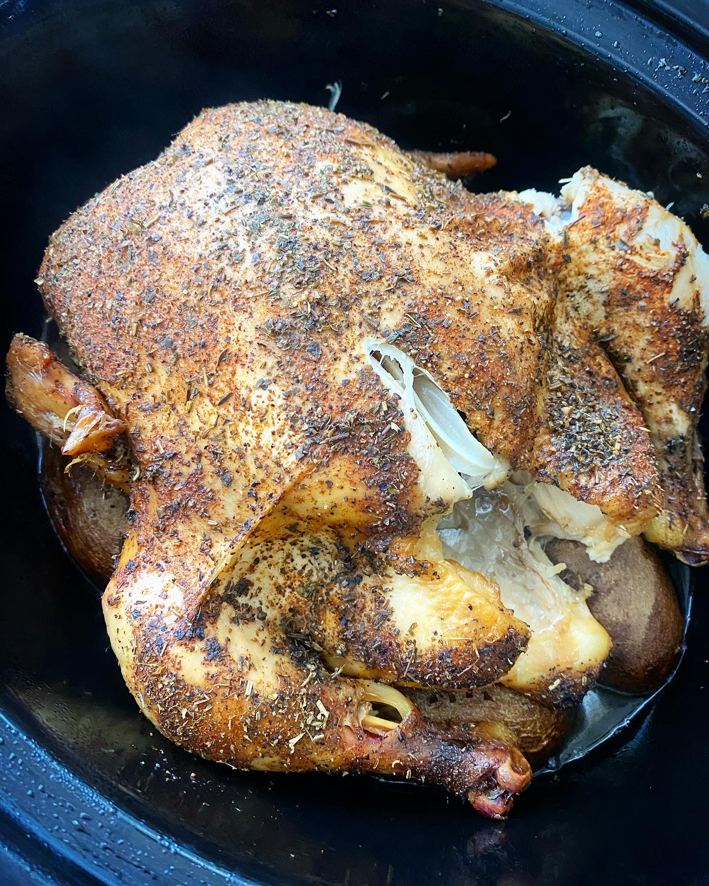 overhead shot of cooked whole chicken on top of potatoes in a black slow cooker