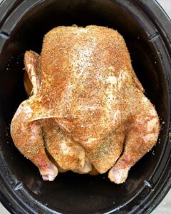 overhead shot of uncooked whole chicken on top of potatoes in a black slow cooker