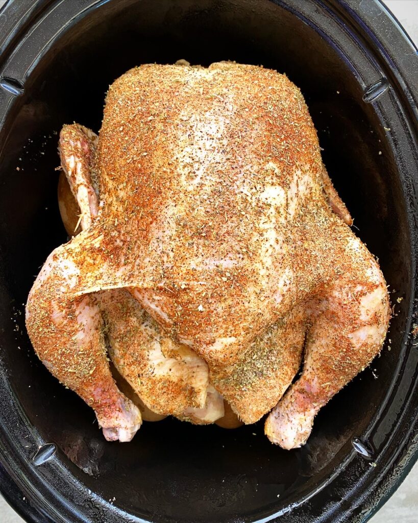 overhead shot of uncooked whole chicken on top of potatoes in a black slow cooker