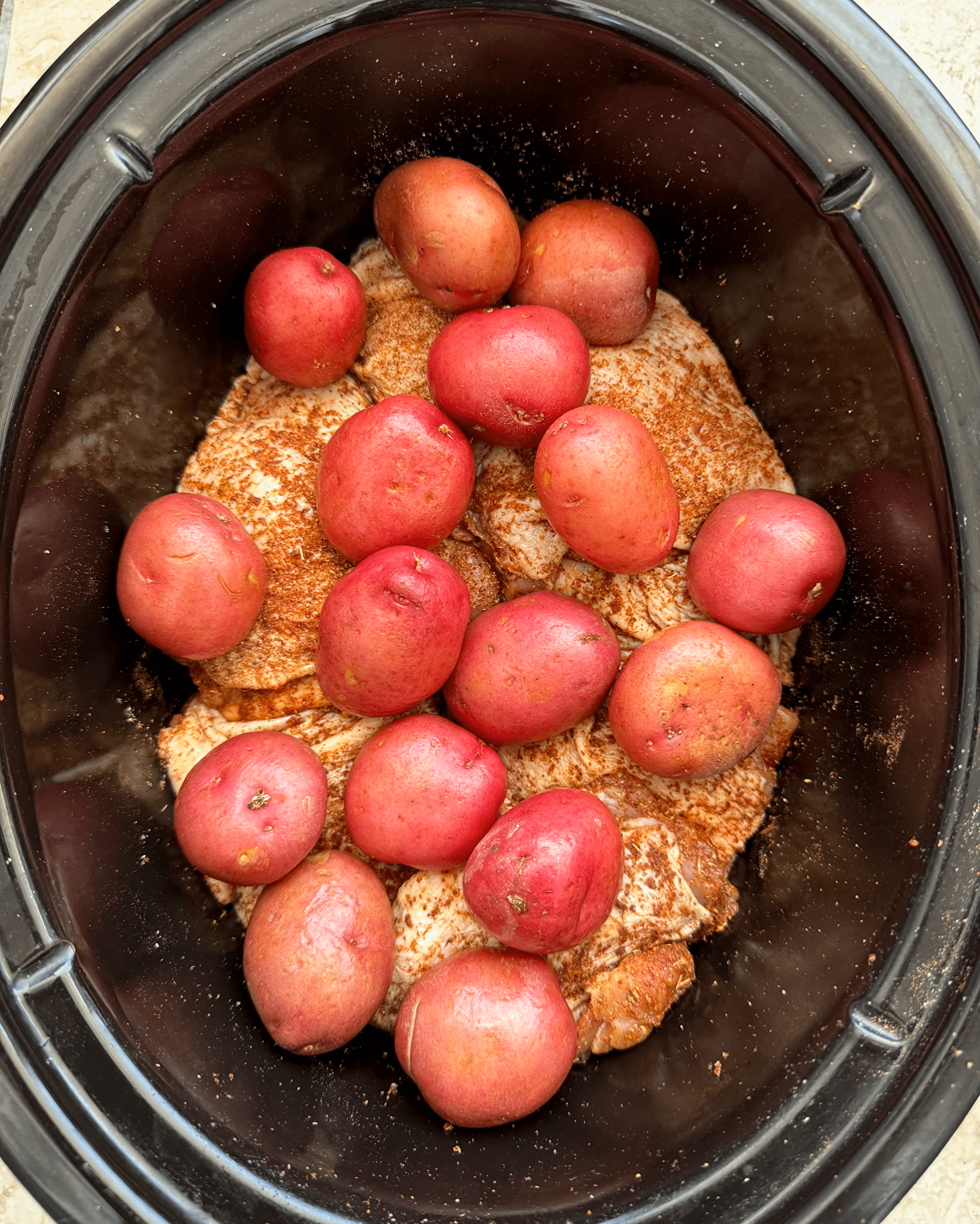 overhead shot of uncooked chicken thigh and potatoes in a black slow cooker