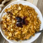 side, close up view of pumpkin oatmeal in a white bowl with raisins on top, almond milk, and a silver spoon