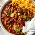 cooked turkey chili in a white bowl topped with cheese, green onion and sour cream with a silver spoon