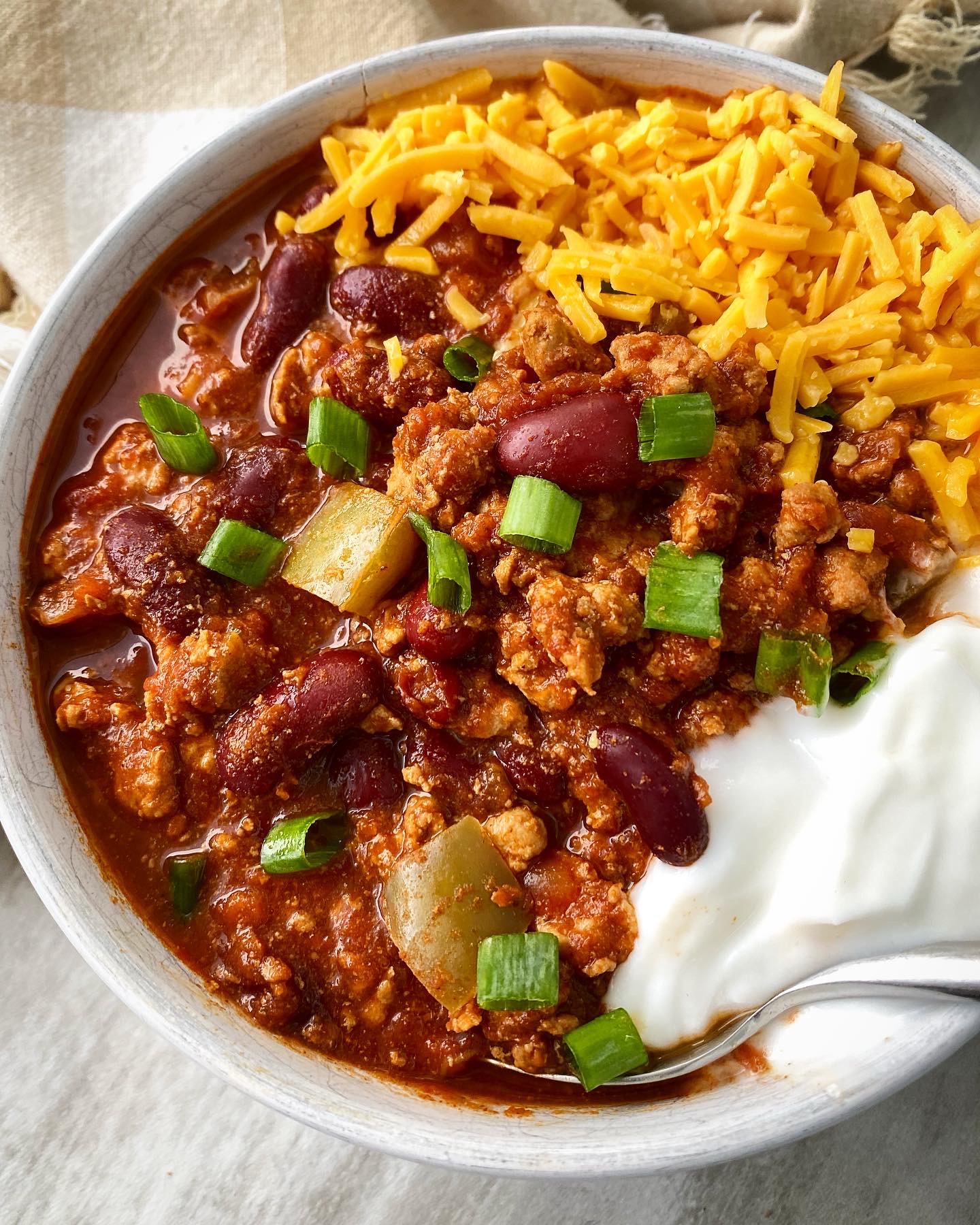 cooked turkey chili in a white bowl topped with cheese, green onion and sour cream with a silver spoon
