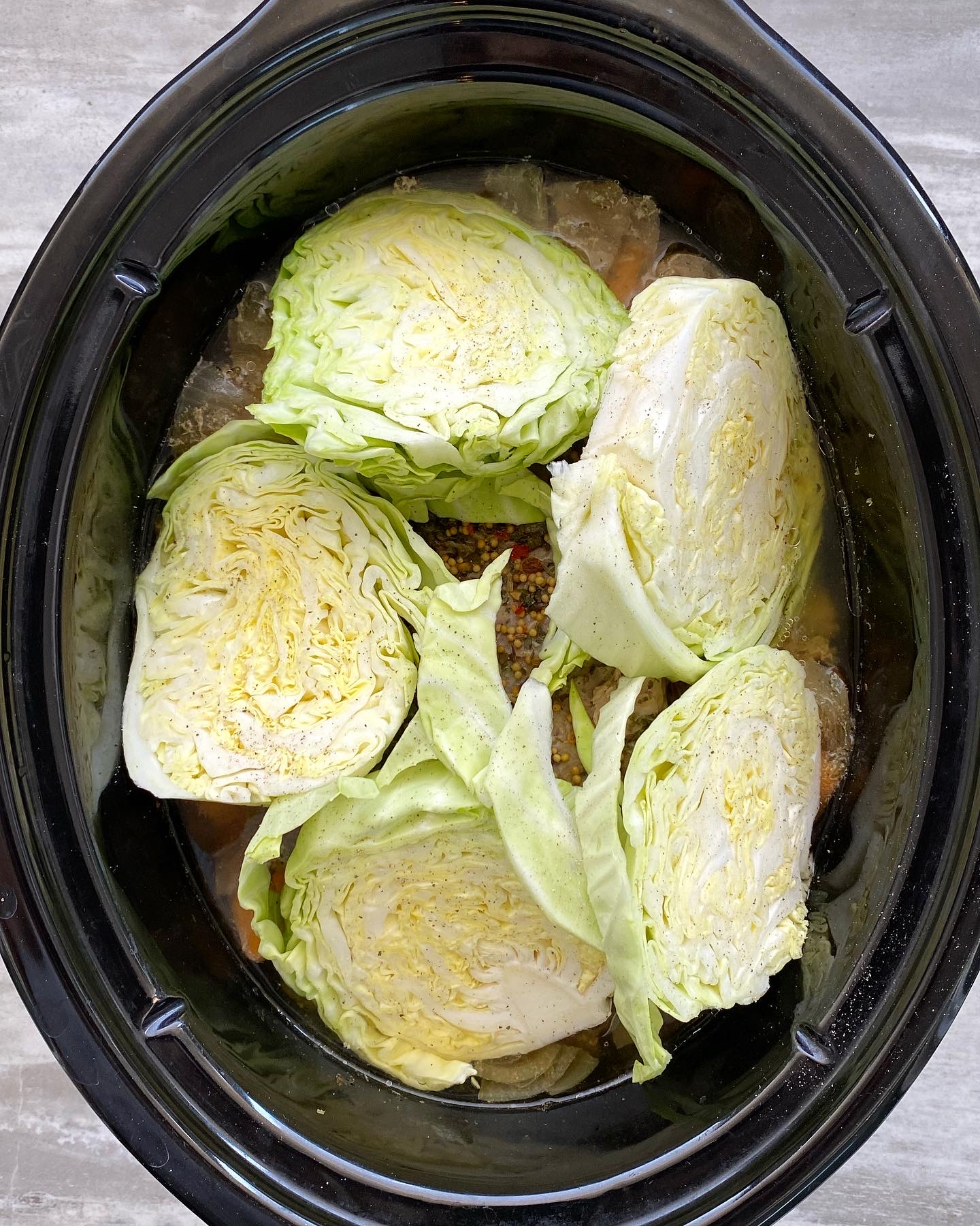 overhead shot of cooked corned beef with potatoes, carrots, and onion in a black slow cooker with uncooked cabbage wedges on top