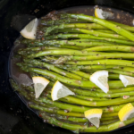 close up shot of Slow Cooker Lemon Garlic Asparagus in a black slow cooker, garnished with chopped lemon