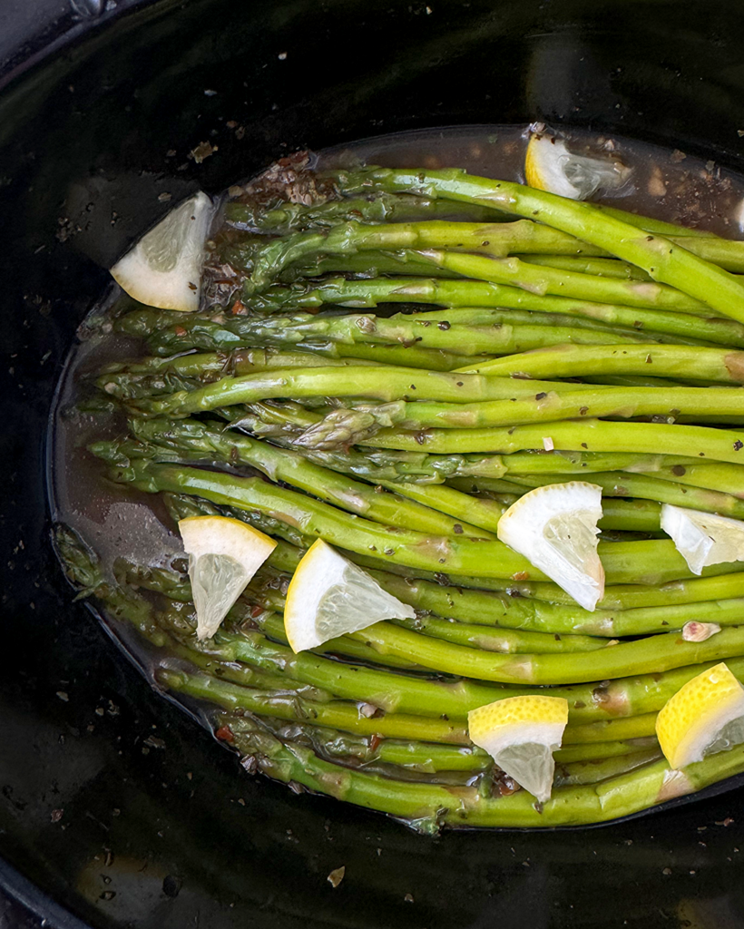 close up shot of Slow Cooker Lemon Garlic Asparagus in a black slow cooker, garnished with chopped lemon
