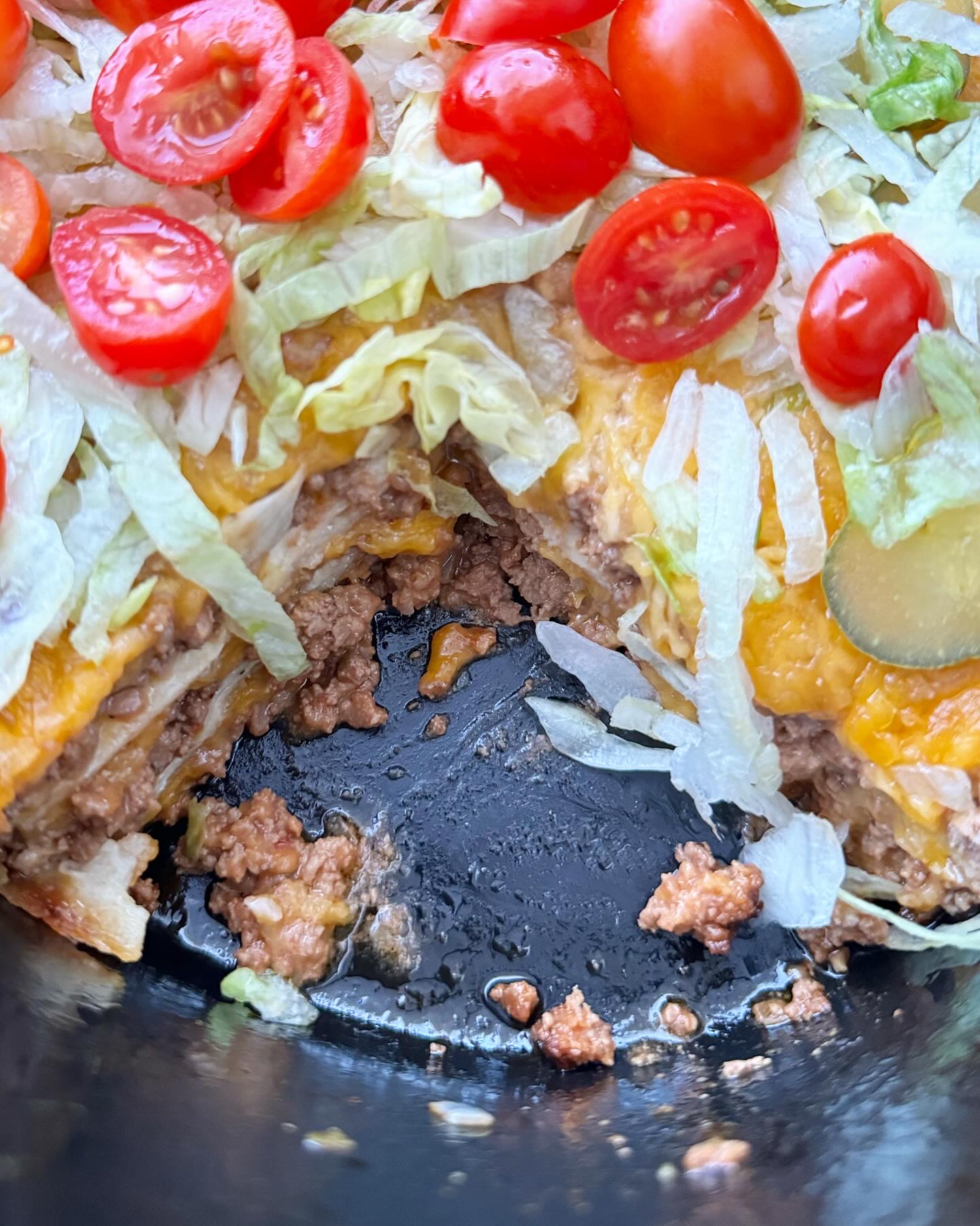 overhead shot of cooked cheeseburger casserole in a black slow cooker garnished with shredded lettuce, pickles, and diced tomatoes with a large slice missing