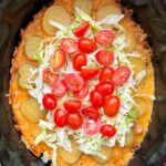 overhead shot of cooked cheeseburger casserole in a black slow cooker garnished with shredded lettuce, pickles, and diced tomatoes
