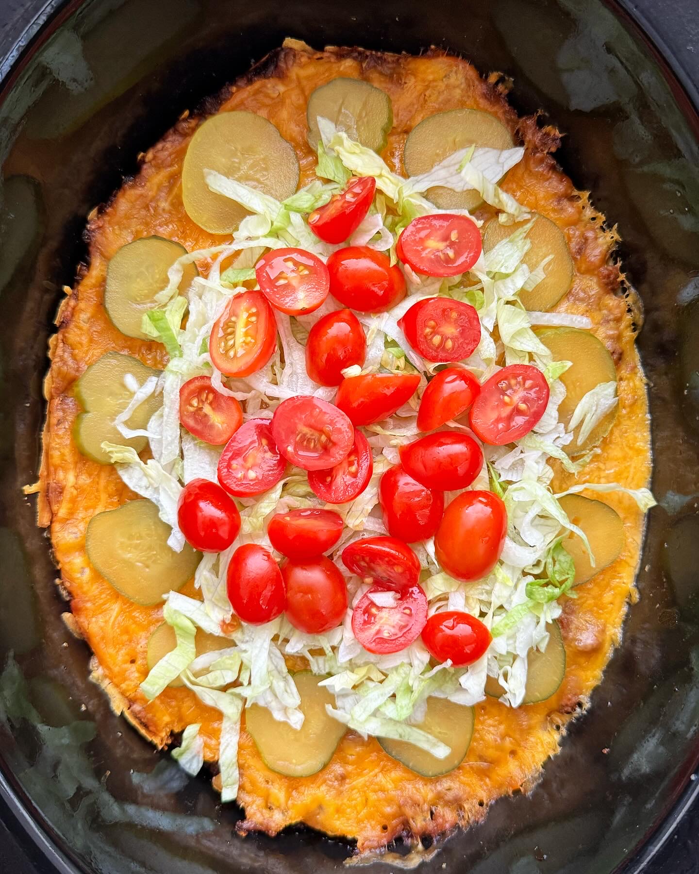 overhead shot of cooked cheeseburger casserole in a black slow cooker garnished with shredded lettuce, pickles, and diced tomatoes