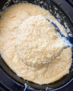 overhead shot of cooked grits in a black slow cooker with a black spoon