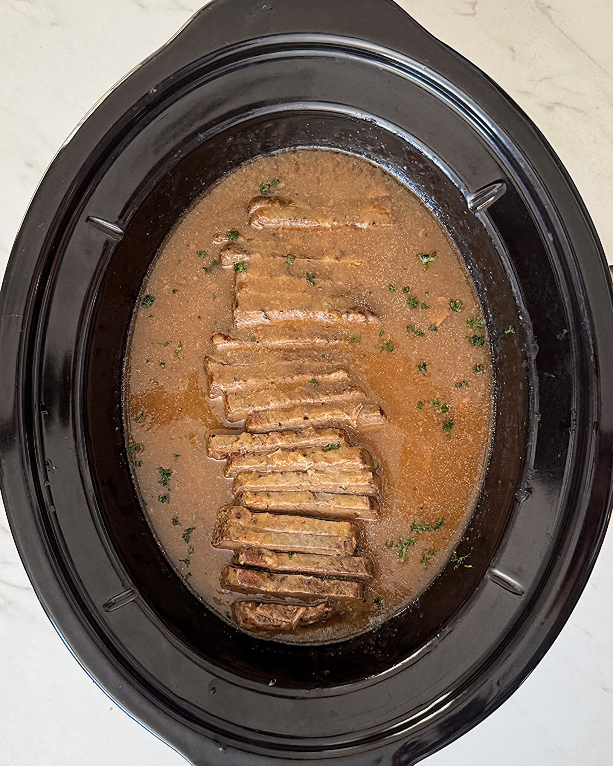overhead shot of cooked brisket and gravy in a black slow cooker that has been sliced