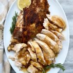 overhead shot of half turkey breast with skin and half sliced turkey breast. Garnished with 1 slice of lemon and fresh rosemary