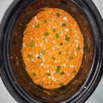 overhead shot of Buffalo Chicken Dip in a black slow cooker garnished with green onions and blue cheese crumbles