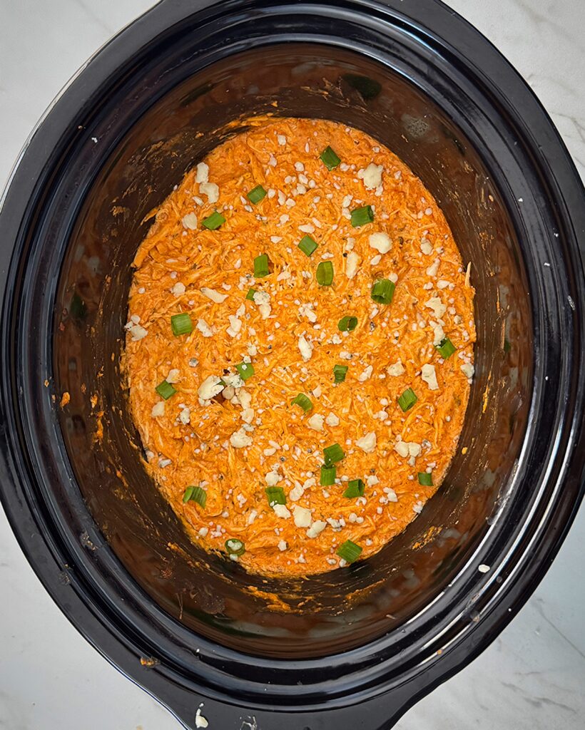 overhead shot of Buffalo Chicken Dip in a black slow cooker garnished with green onions and blue cheese crumbles