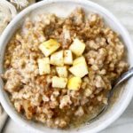 overhead shot of a slow cooker apple cinnamon oatmeal in a white bowl with diced apple on top & milk with a silver spoon