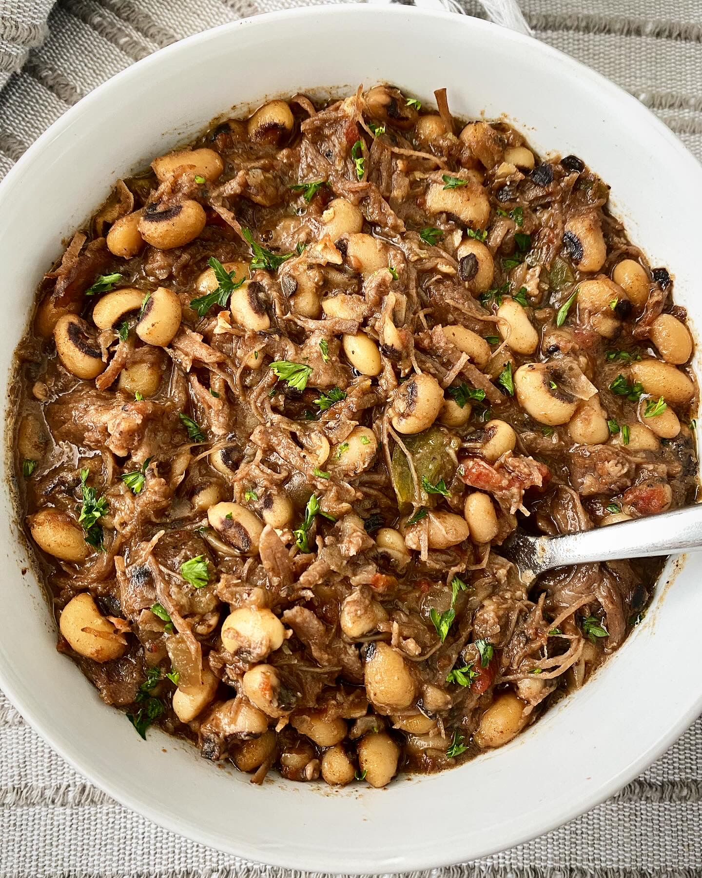 close up shot of cooked shredded beef and black eyed peas in a white bowl with a silver spoon
