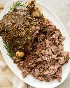 overhead shot of cooked leg of lamb on a white platter, half shredded