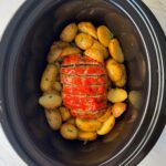 overhead shot of cooked, sliced meatloaf in a black slow cooker surrounded by potatoes