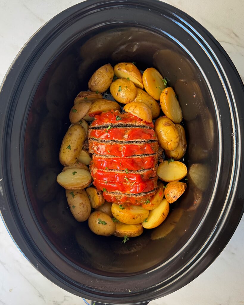 overhead shot of cooked, sliced meatloaf in a black slow cooker surrounded by potatoes