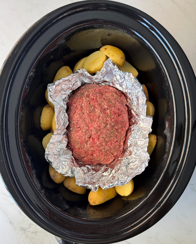 overhead shot of uncooked meatloaf and potatoes in a black slow cooker