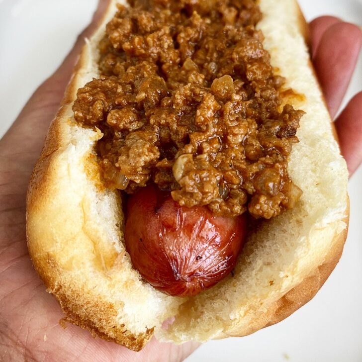 close up shot of a hot dog with chili on top held in a black left hand