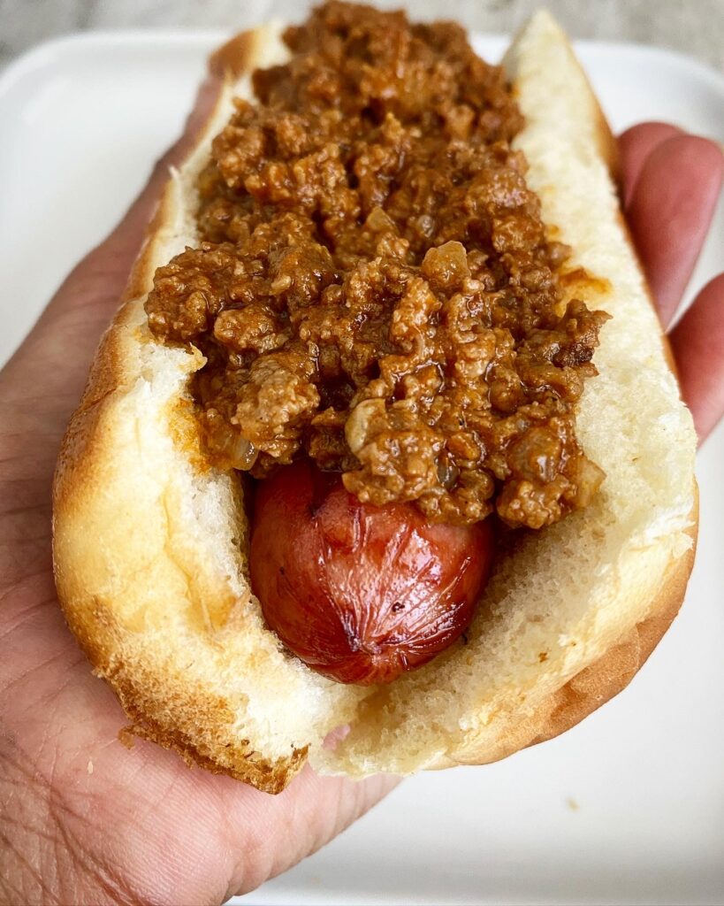 close up shot of a hot dog with chili on top held in a black left hand