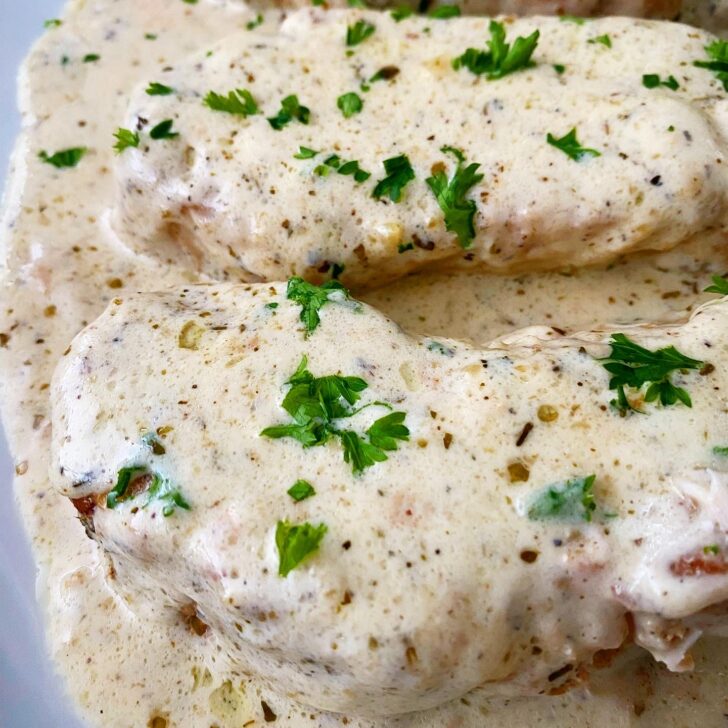 close up, side view shot of 2 pork chops on a white plate with a cream sauce on top, garnished with chopped fresh parsley