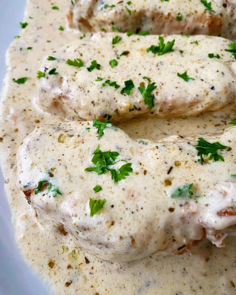 close up, side view shot of 2 pork chops on a white plate with a cream sauce on top, garnished with chopped fresh parsley