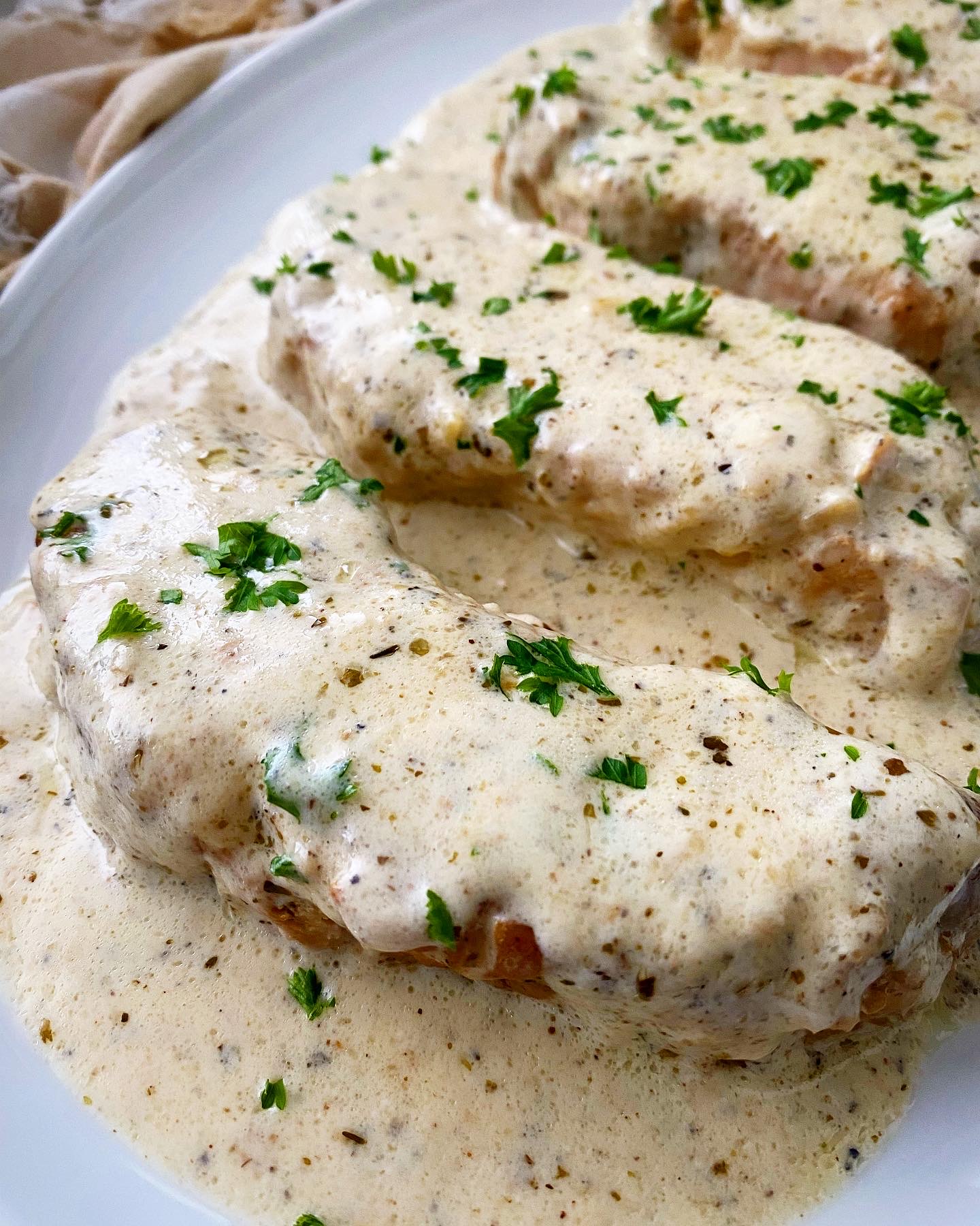 close up, side view shot of 4 cooked pork chops on a white plate with a cream sauce on top, garnished with chopped fresh parsley